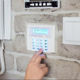 Elektrokol - Dłoń wciskająca przycisk na białym panelu alarmowym z niebieskim podświetleniem, zamontowanym na ścianie z cegły dekoracyjnej, obok biały domofon i włącznik światła.