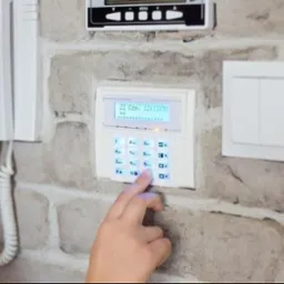 Dłoń wciskająca przycisk na białym panelu alarmowym z niebieskim podświetleniem, zamontowanym na ścianie z cegły dekoracyjnej, obok biały domofon i włącznik światła.