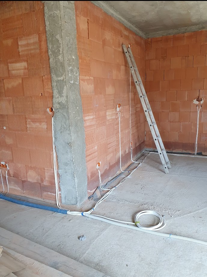 Wnętrze w trakcie prac wykończeniowych: ściany z cegły dziurawki, surowy betonowy filar, drabina oparta o ścianę, widoczne przewody elektryczne wychodzące z puszek instalacyjnych, niebieska rura...