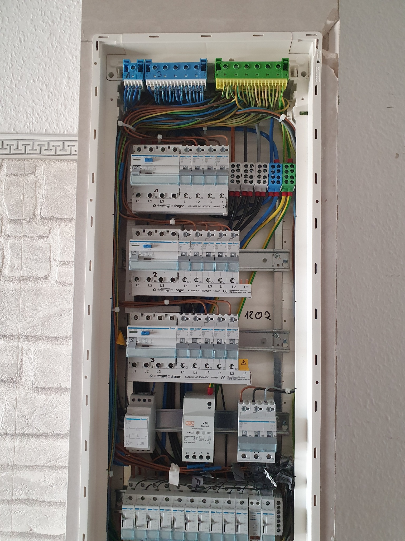 Otwarta skrzynka z zabezpieczeniami elektrycznymi Hager, widoczne różnokolorowe przewody, bezpieczniki i listwy zaciskowe na tle fragmentu otynkowanej ściany.