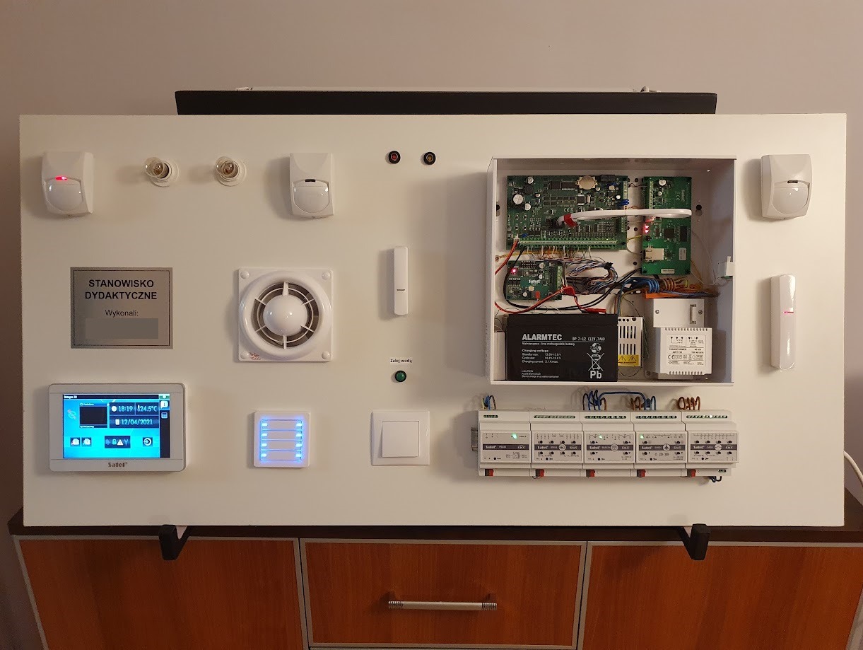 Panel demonstracyjny systemów zabezpieczeń: czujniki ruchu, centrala alarmowa z akumulatorem, panel sterowania dotykowy, sygnalizator wewnętrzny, elementy instalacji elektrycznej, wentylator...