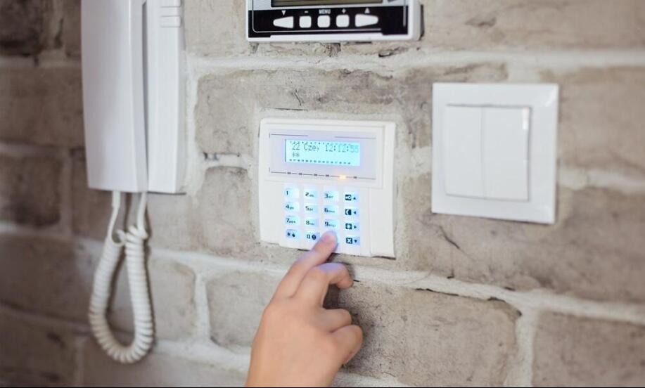 Dłoń wciskająca przycisk na białym panelu alarmowym z niebieskim podświetleniem, zamontowanym na ścianie z cegły dekoracyjnej, obok biały domofon i włącznik światła.