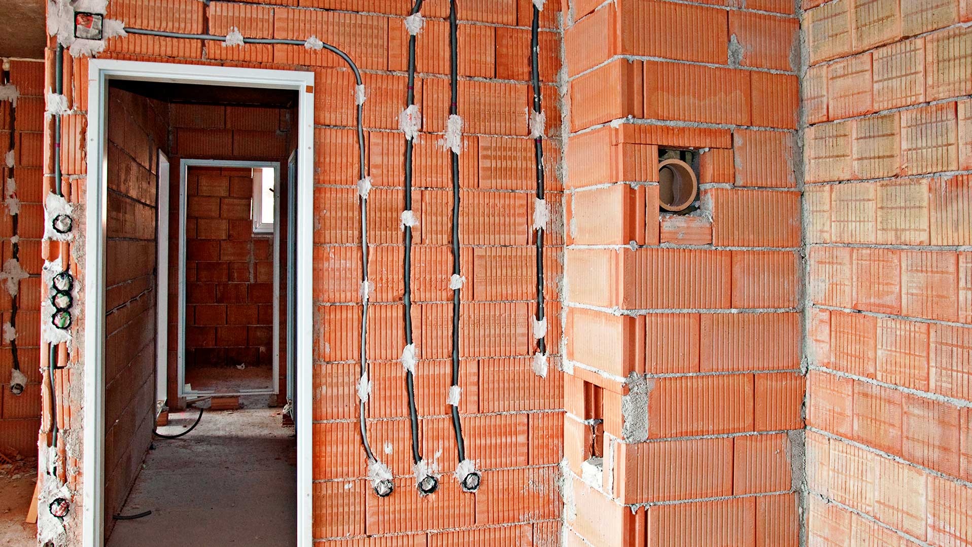 Surowe wnętrze w trakcie budowy z widocznymi przewodami elektrycznymi przymocowanymi do ściany z cegieł dziurawki za pomocą gipsu, otwory pod gniazdka oraz widok na kolejne pomieszczenia przez...