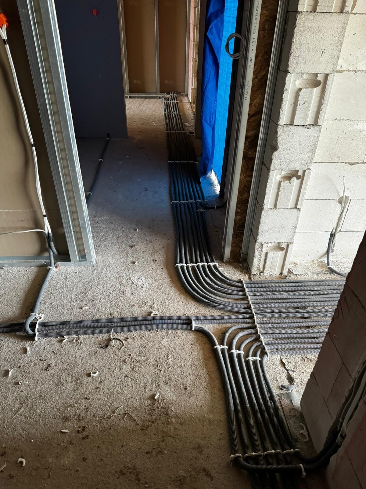 Szczegółowe ułożenie peszli z kablami elektrycznymi na surowym podłożu w trakcie remontu, widoczne ściany z bloczków i profile metalowe.