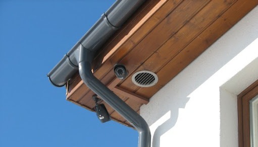 Kamera monitoringu zamontowana pod okapem drewnianej podbitki dachowej, obok kratki wentylacyjnej i szarej rynny spustowej na tle błękitnego nieba.