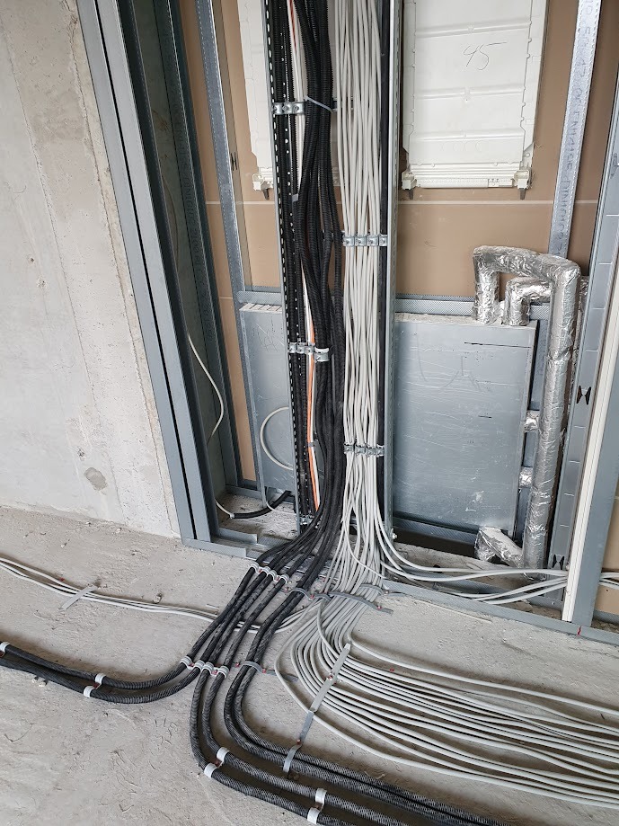 Instalacja elektryczna w trakcie montażu w surowym stanie, widoczne przewody w peszlach, kable białe, czarne i pomarańczowe, metalowe profile konstrukcyjne ścian i puszka instalacyjna.