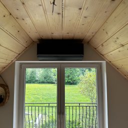 AMPERE SPÓŁKA Z OGRANICZONĄ ODPOWIEDZIALNOŚCIĄ - Czarny klimatyzator zamontowany pod drewnianym sufitem na poddaszu, widok na zielony krajobraz przez balkonowe okno.