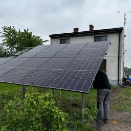 AMPERE SPÓŁKA Z OGRANICZONĄ ODPOWIEDZIALNOŚCIĄ - Montaż paneli fotowoltaicznych na metalowej konstrukcji przed domem jednorodzinnym z anteną na dachu, widoczny instalator w szarych spodniach roboczych.
