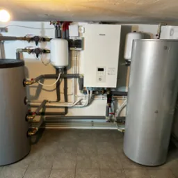 Zestaw dwóch szarych zbiorników akumulacyjnych ciepła, białego kotła grzewczego Panasonic i białych zbiorników wyrównawczych w kotłowni z widocznymi rurami i zaworami.