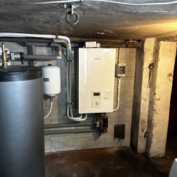 AMPERE SPÓŁKA Z OGRANICZONĄ ODPOWIEDZIALNOŚCIĄ - Biały wiszący piec gazowy marki Panasonic w kotłowni, obok szary zbiornik na wodę i biały zbiornik wyrównawczy, instalacja rur grzewczych pod sufitem, surowy betonowy sufit i kolumna.