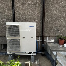 AMPERE SPÓŁKA Z OGRANICZONĄ ODPOWIEDZIALNOŚCIĄ - Zewnętrzna jednostka pompy ciepła Panasonic zainstalowana przy ścianie budynku z widocznymi rurami i roślinami w donicach w tle.