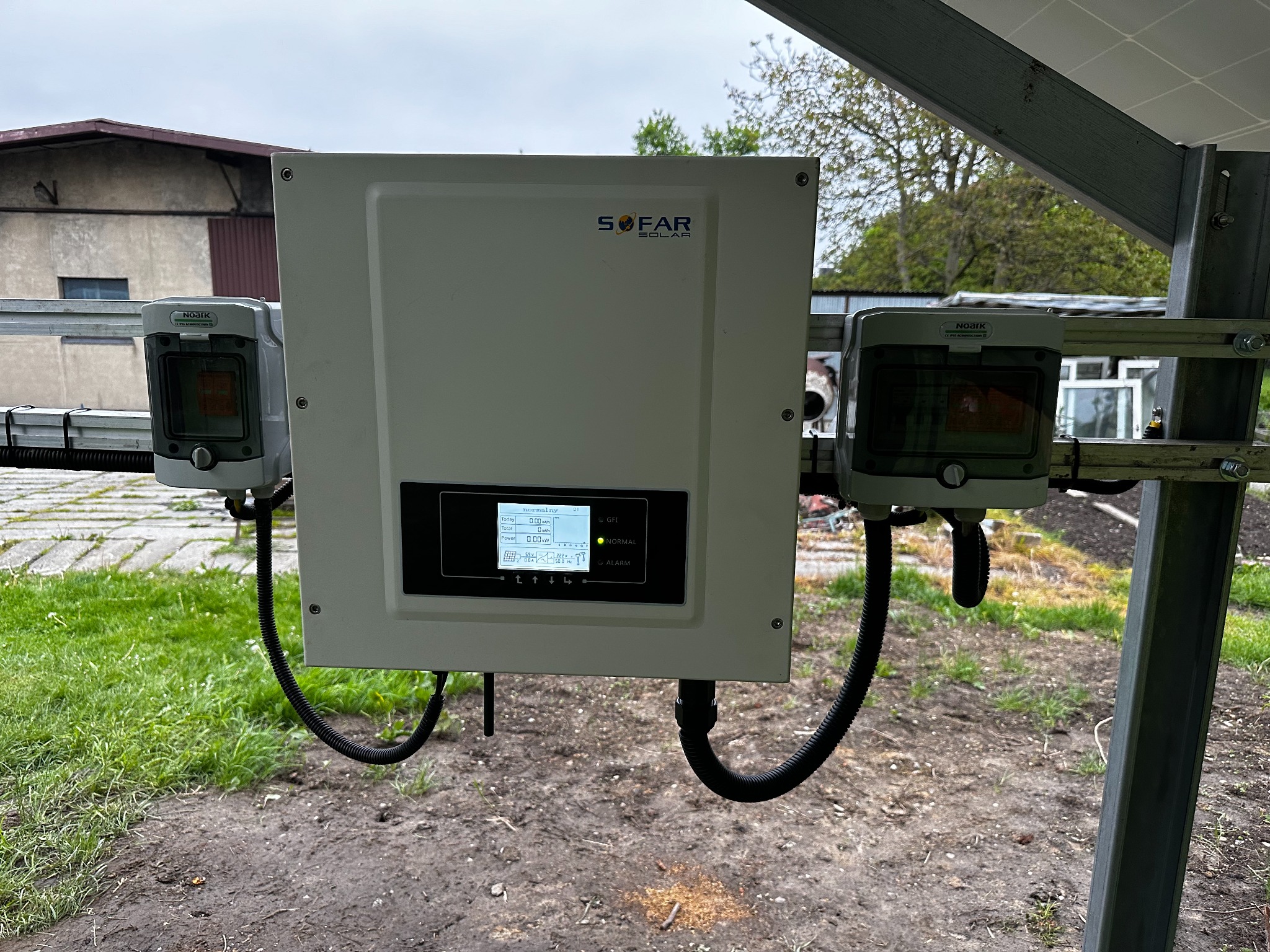 Biały falownik Sofar Solar z wyświetlaczem i czarnymi kablami, zamontowany na konstrukcji wsporczej paneli fotowoltaicznych, z dwoma szarymi skrzynkami Noark po bokach, na tle budynku i drzew.