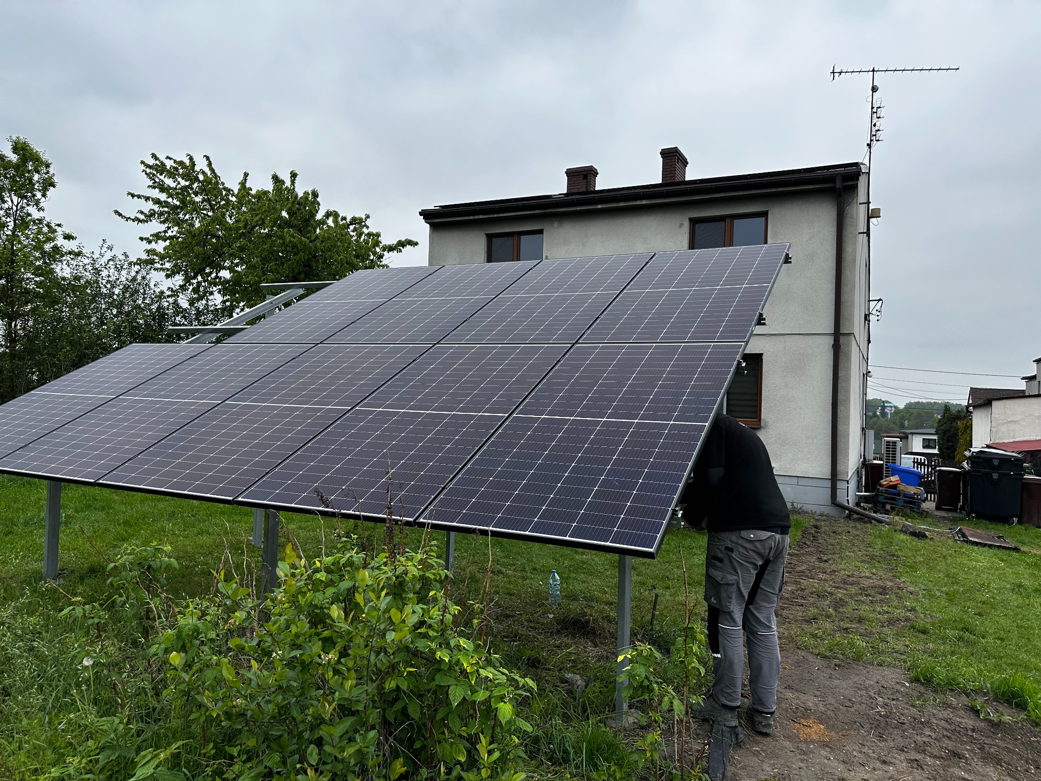 Montaż paneli fotowoltaicznych na metalowej konstrukcji przed domem jednorodzinnym z anteną na dachu, widoczny instalator w szarych spodniach roboczych.
