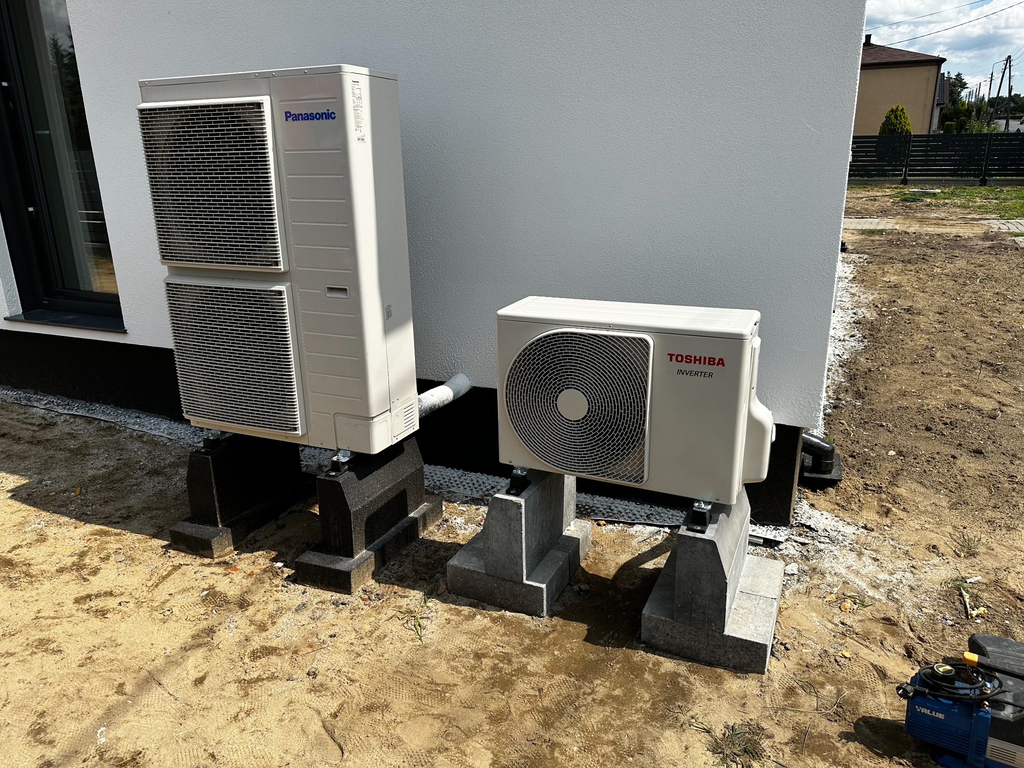 Dwie zewnętrzne jednostki klimatyzacji: Panasonic ustawiona pionowo i Toshiba Inverter, obie na betonowych podstawach przy białej ścianie budynku, widoczna pompa próżniowa w rogu.
