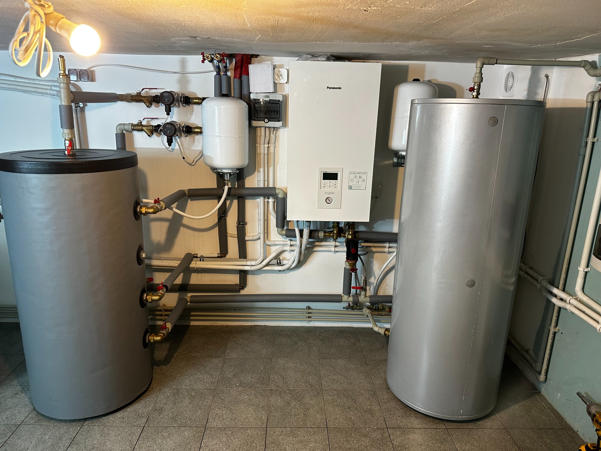 Zestaw dwóch szarych zbiorników akumulacyjnych ciepła, białego kotła grzewczego Panasonic i białych zbiorników wyrównawczych w kotłowni z widocznymi rurami i zaworami.