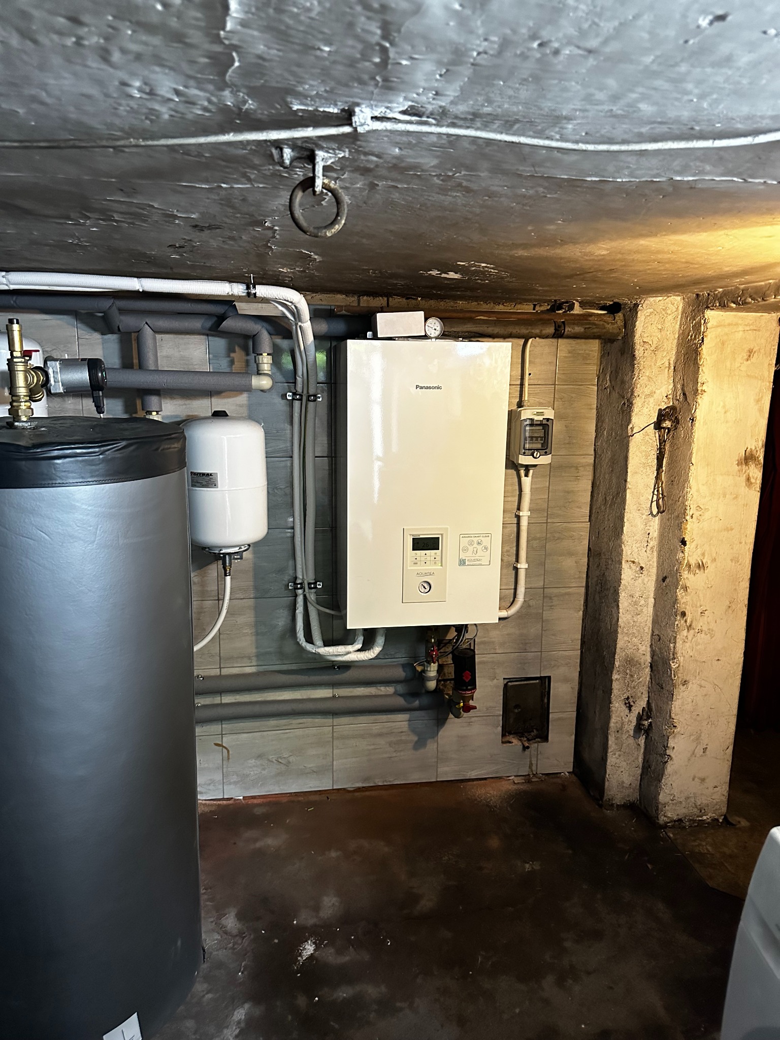 Biały wiszący piec gazowy marki Panasonic w kotłowni, obok szary zbiornik na wodę i biały zbiornik wyrównawczy, instalacja rur grzewczych pod sufitem, surowy betonowy sufit i kolumna.