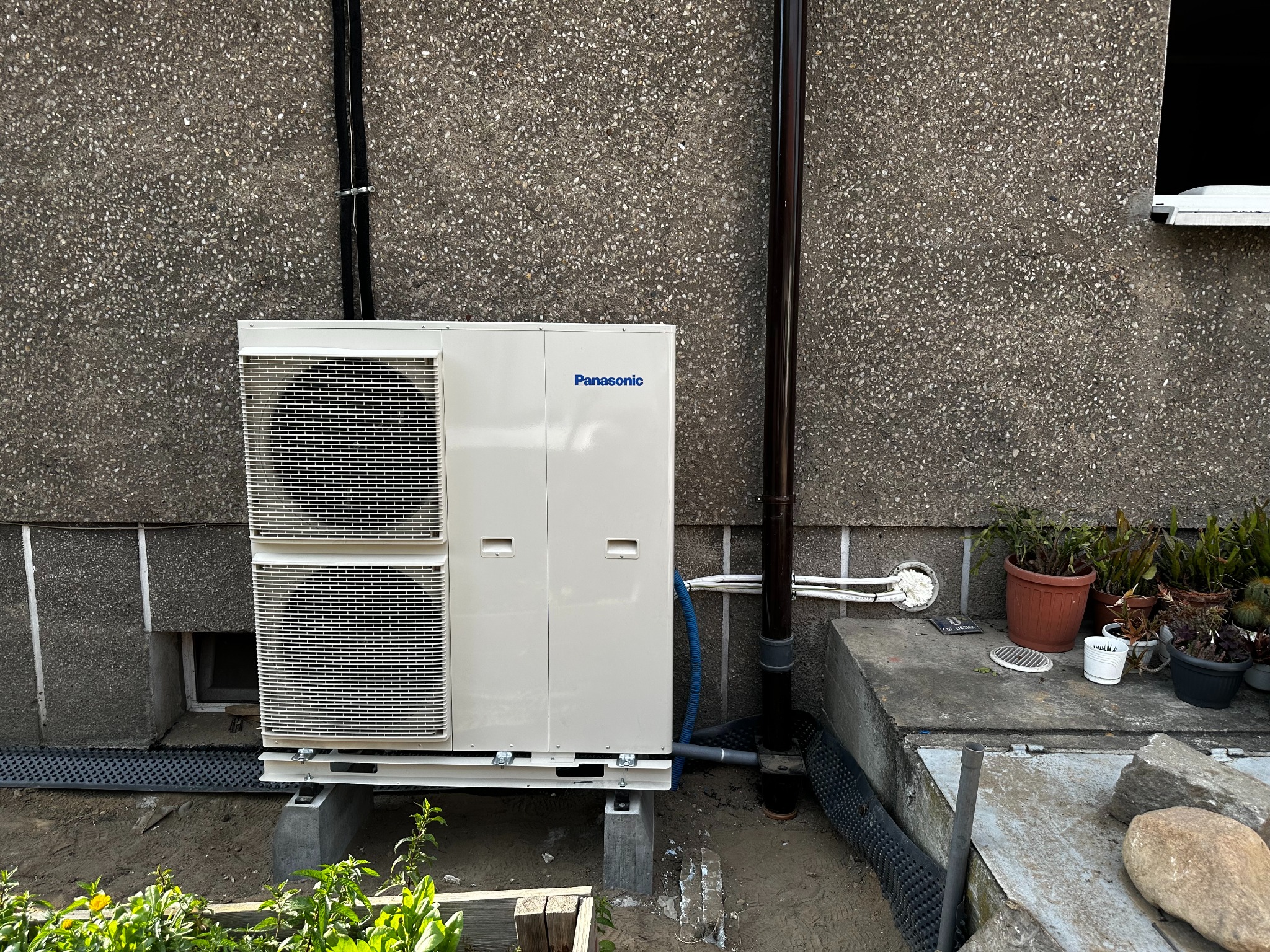 Zewnętrzna jednostka pompy ciepła Panasonic zainstalowana przy ścianie budynku z widocznymi rurami i roślinami w donicach w tle.