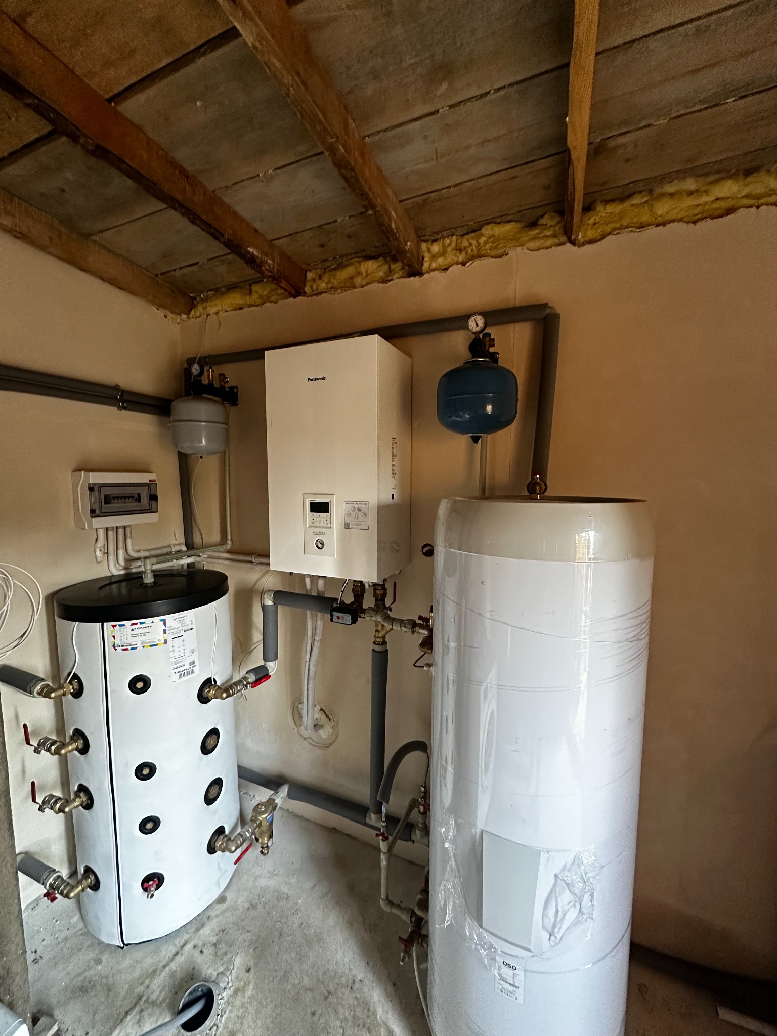 Zestaw urządzeń do ogrzewania wody i centralnego ogrzewania w kotłowni: biały zasobnik ciepłej wody owinięty folią, biały bojler z czarnymi otworami i licznymi zaworami, wiszący na ścianie biały...