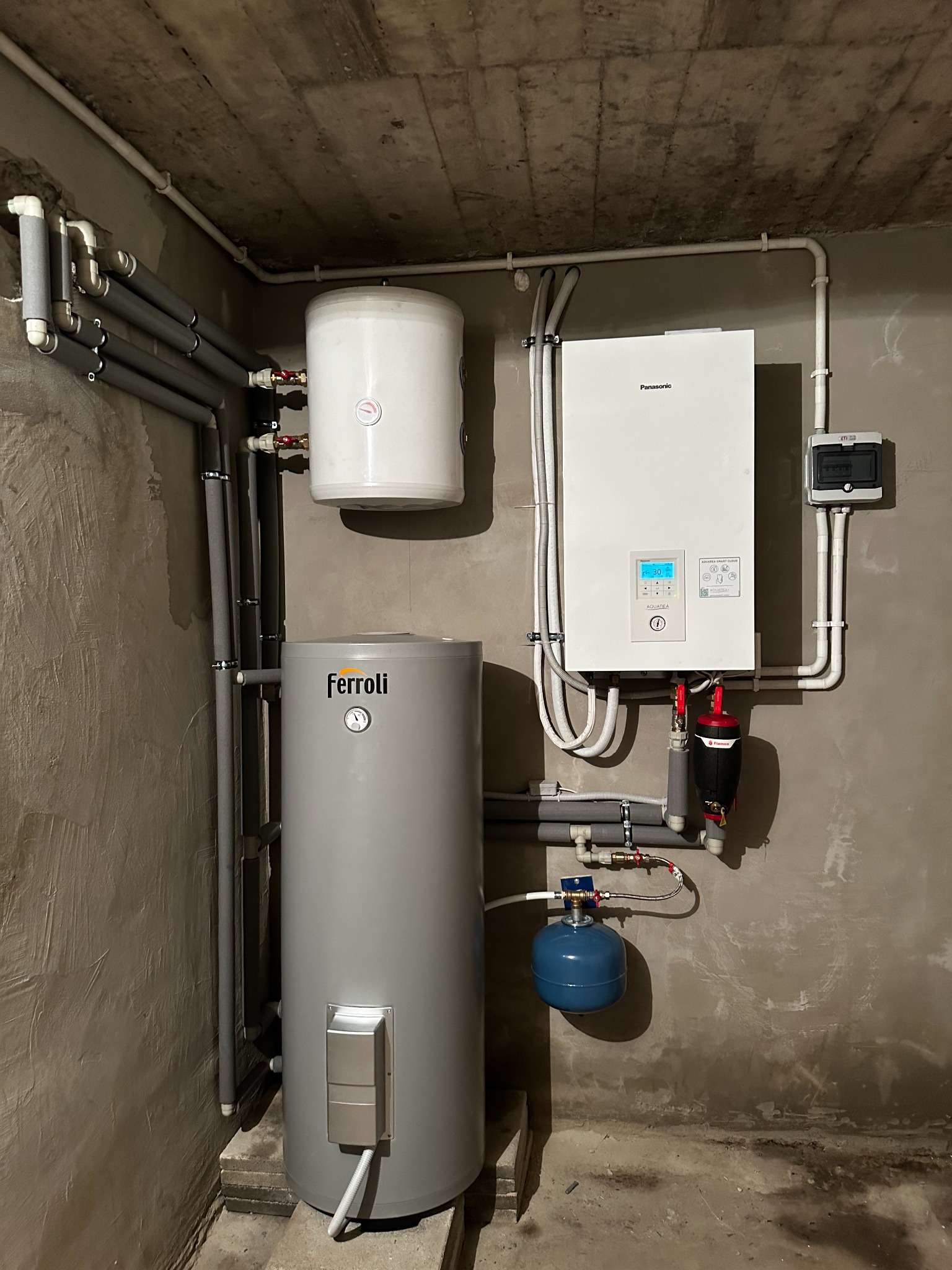 Instalacja grzewcza w surowym pomieszczeniu: pionowy szary zbiornik Ferroli z manometrem, biały zbiornik akumulacyjny, wiszący biały moduł Panasonic z wyświetlaczem cyfrowym oraz liczne rury...