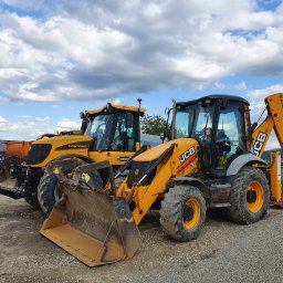 Andrzej Obrzut - Dwie żółte maszyny budowlane JCB i Fastrac na żwirowym podłożu, z widoczną górą w tle i błękitnym niebem z chmurami.