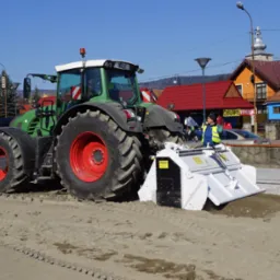 Zielony traktor z czerwonymi felgami i białym urządzeniem do wyrównywania terenu pracuje na piaszczystym placu budowy; w tle budynki mieszkalne i palety z kostką brukową.