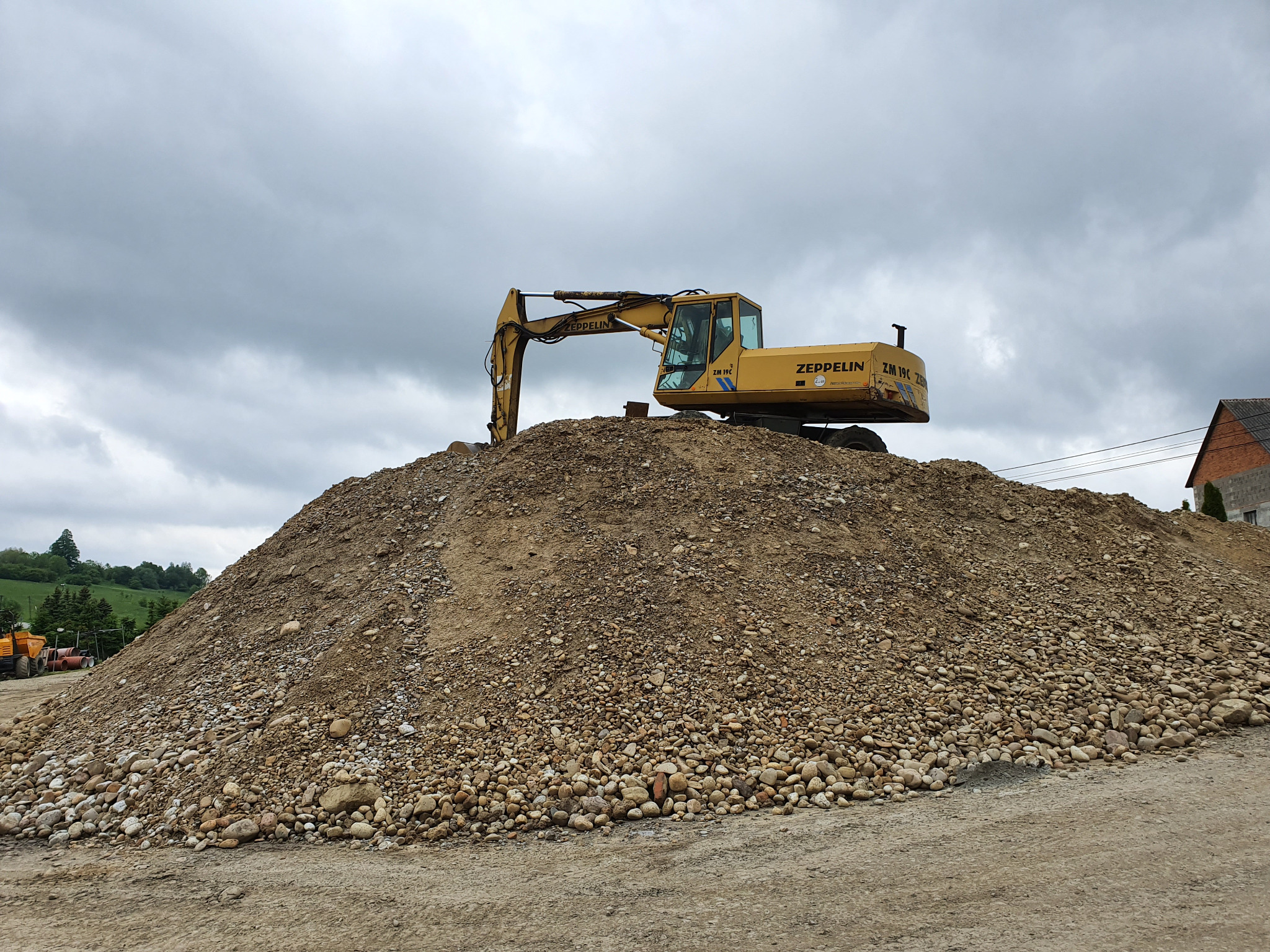 Żółta koparka Zeppelin ZM 19C na szczycie dużej pryzmy ziemi i kamieni, pochmurne niebo w tle.