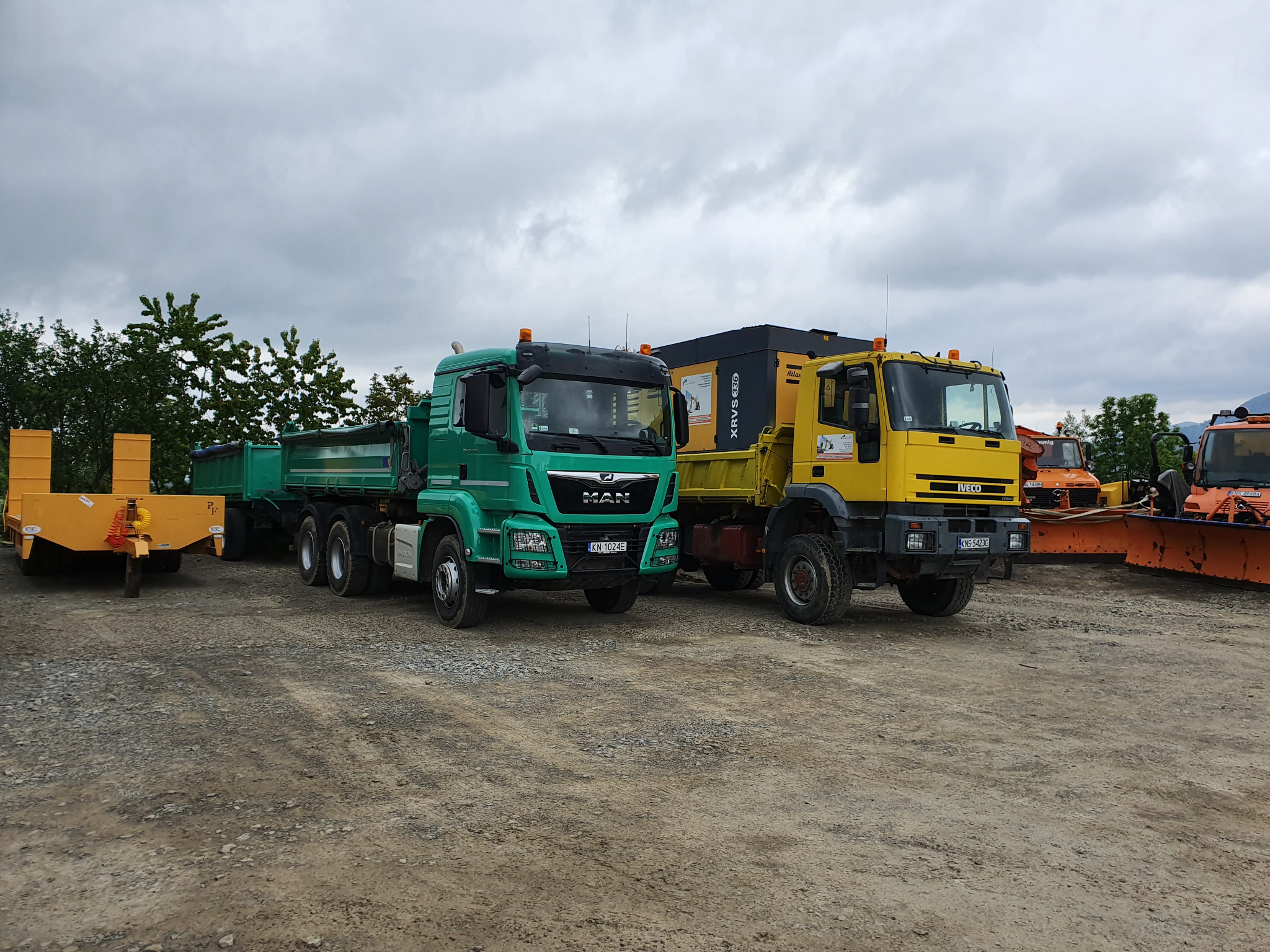 Zielony MAN z przyczepą i żółty IVECO na placu budowy, w tle sprzęt do odśnieżania i pochmurne niebo.