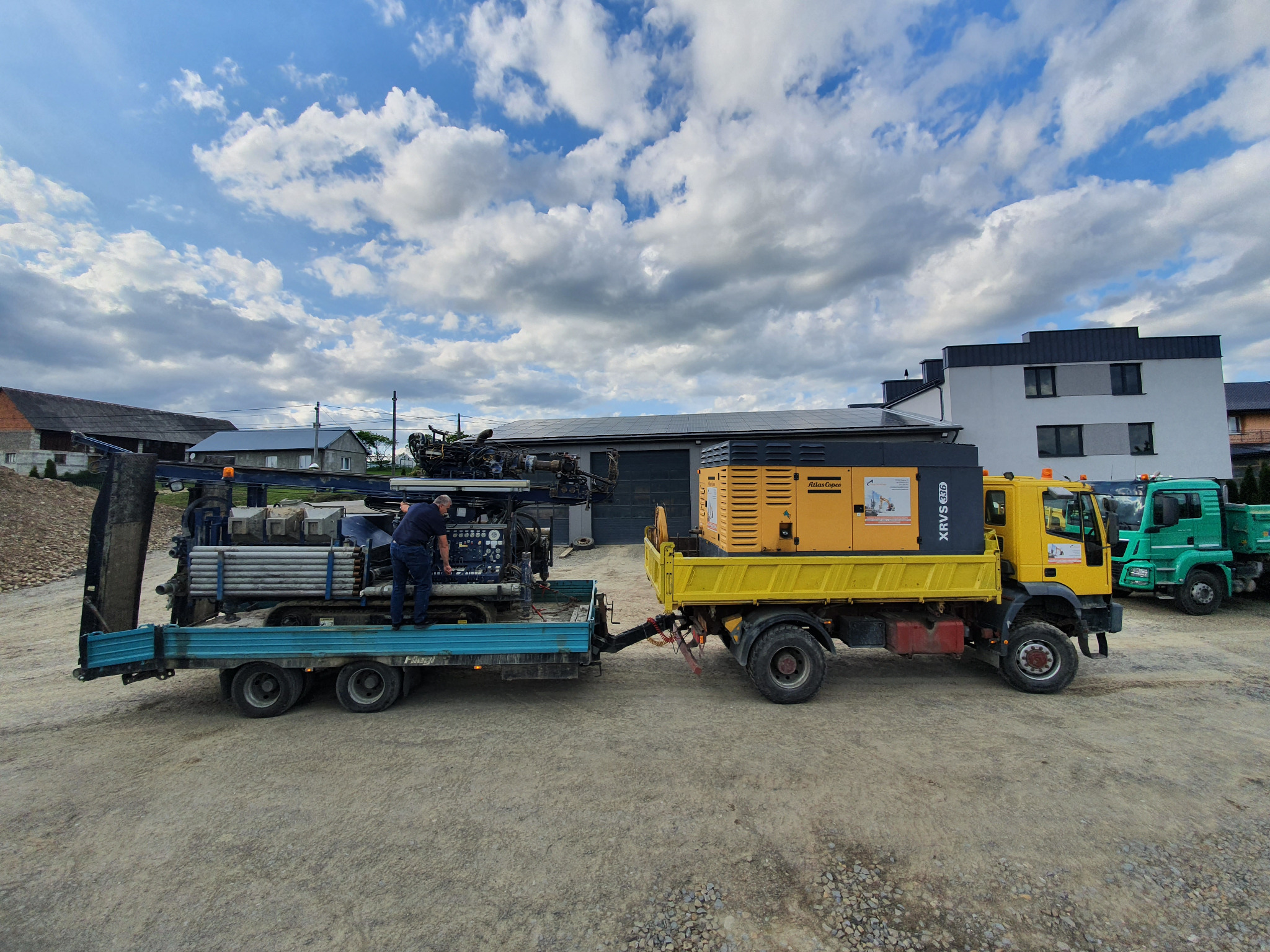 Żółta ciężarówka z generatorem Atlas Copco ciągnie niebieską przyczepę z wiertnicą i rurami wiertniczymi; operator w ciemnej koszuli stoi na przyczepie przy wiertnicy.