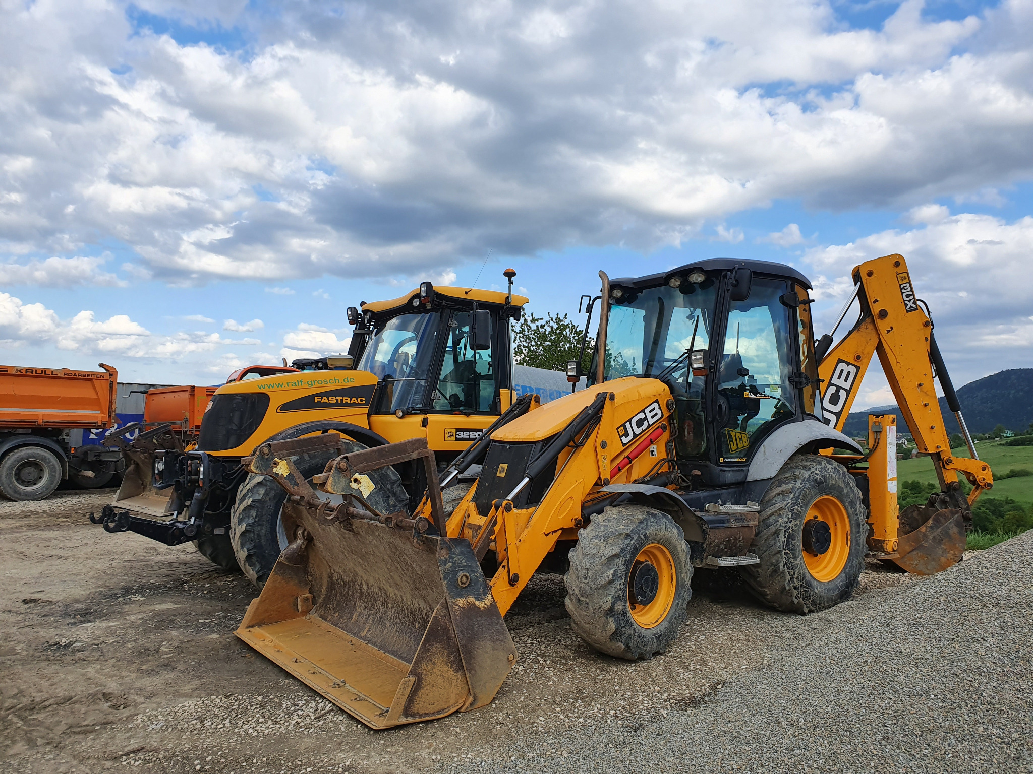 Dwie żółte maszyny budowlane JCB i Fastrac na żwirowym podłożu, z widoczną górą w tle i błękitnym niebem z chmurami.