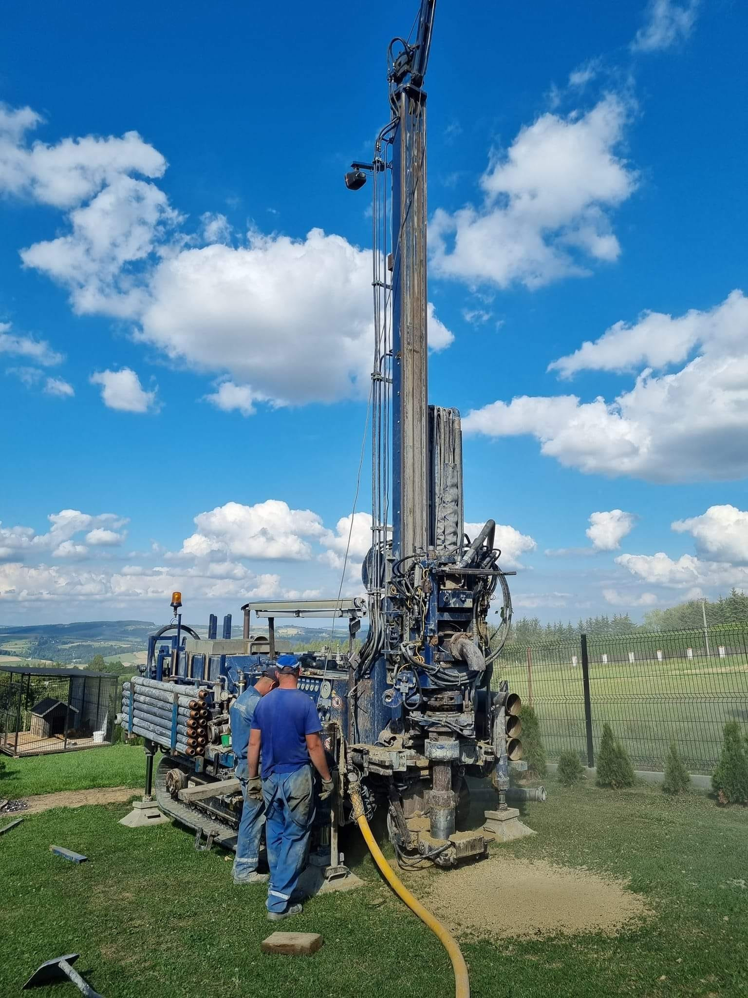 Wiertnica na gąsienicach podczas pracy, dwóch operatorów w kombinezonach, żółty wąż, w tle krajobraz z widokiem na pola i lasy pod błękitnym niebem z białymi chmurami.