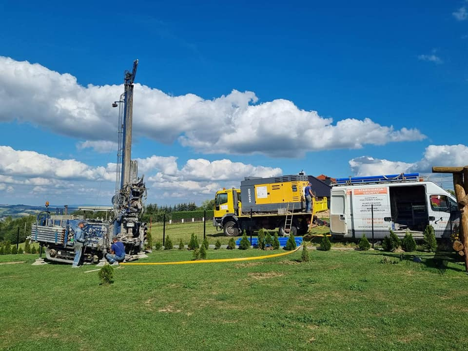 Prace wiertnicze: wiertnica głębinowa na gąsienicach i żółty agregat prądotwórczy na tle zielonego trawnika i błękitnego nieba z chmurami, dwóch pracowników przy maszynie.