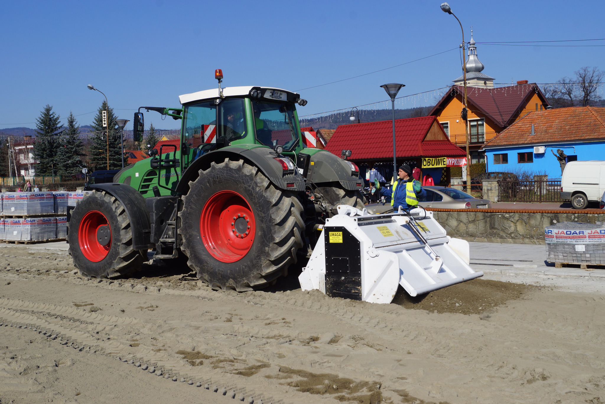 Zielony traktor z czerwonymi felgami i białym urządzeniem do wyrównywania terenu pracuje na piaszczystym placu budowy; w tle budynki mieszkalne i palety z kostką brukową.