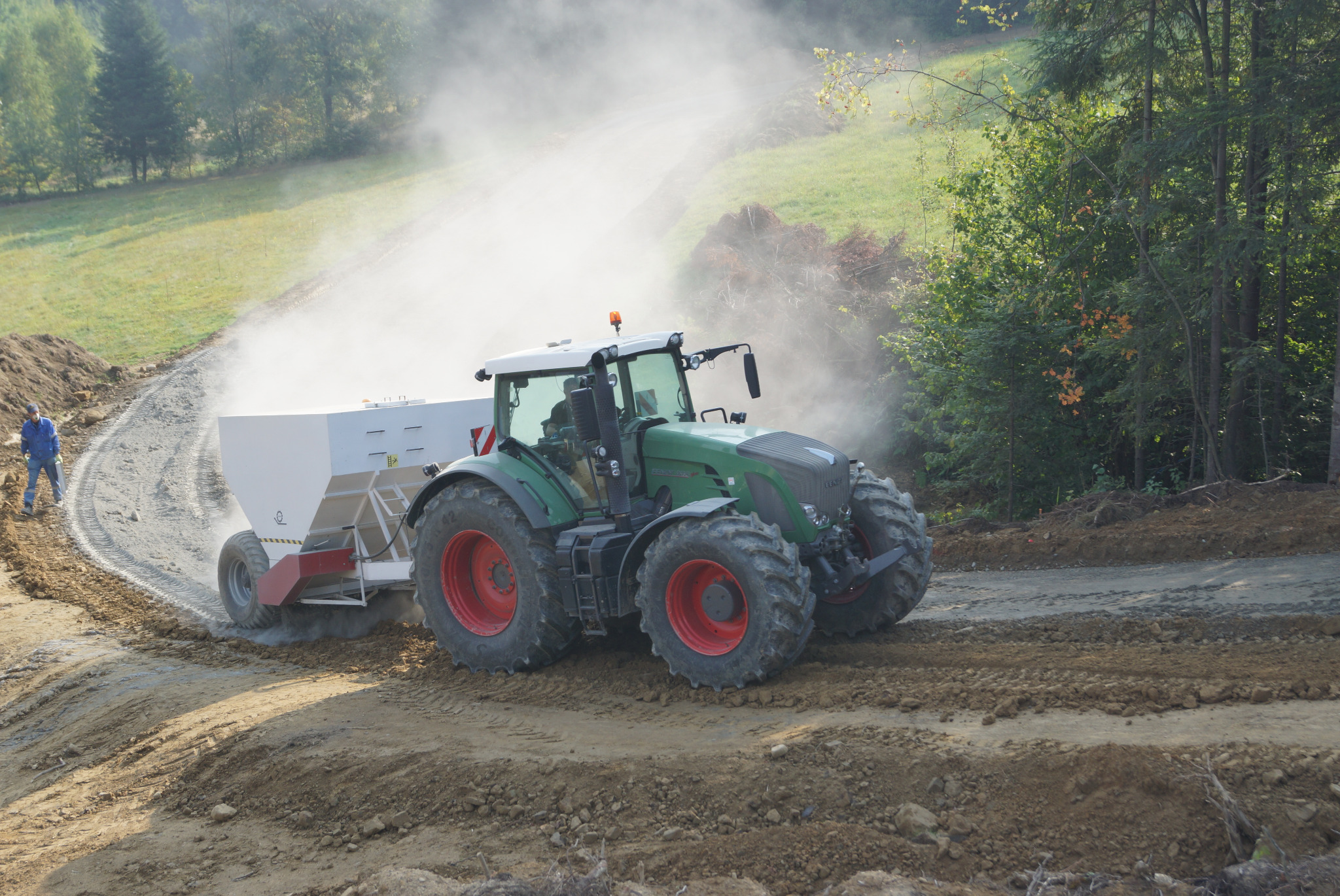 Ciągnik z rozsiewaczem na nierównym terenie w Nowym Sączu, z lewej strony widoczna postać w niebieskim kombinezonie, w tle zalesione wzgórza.