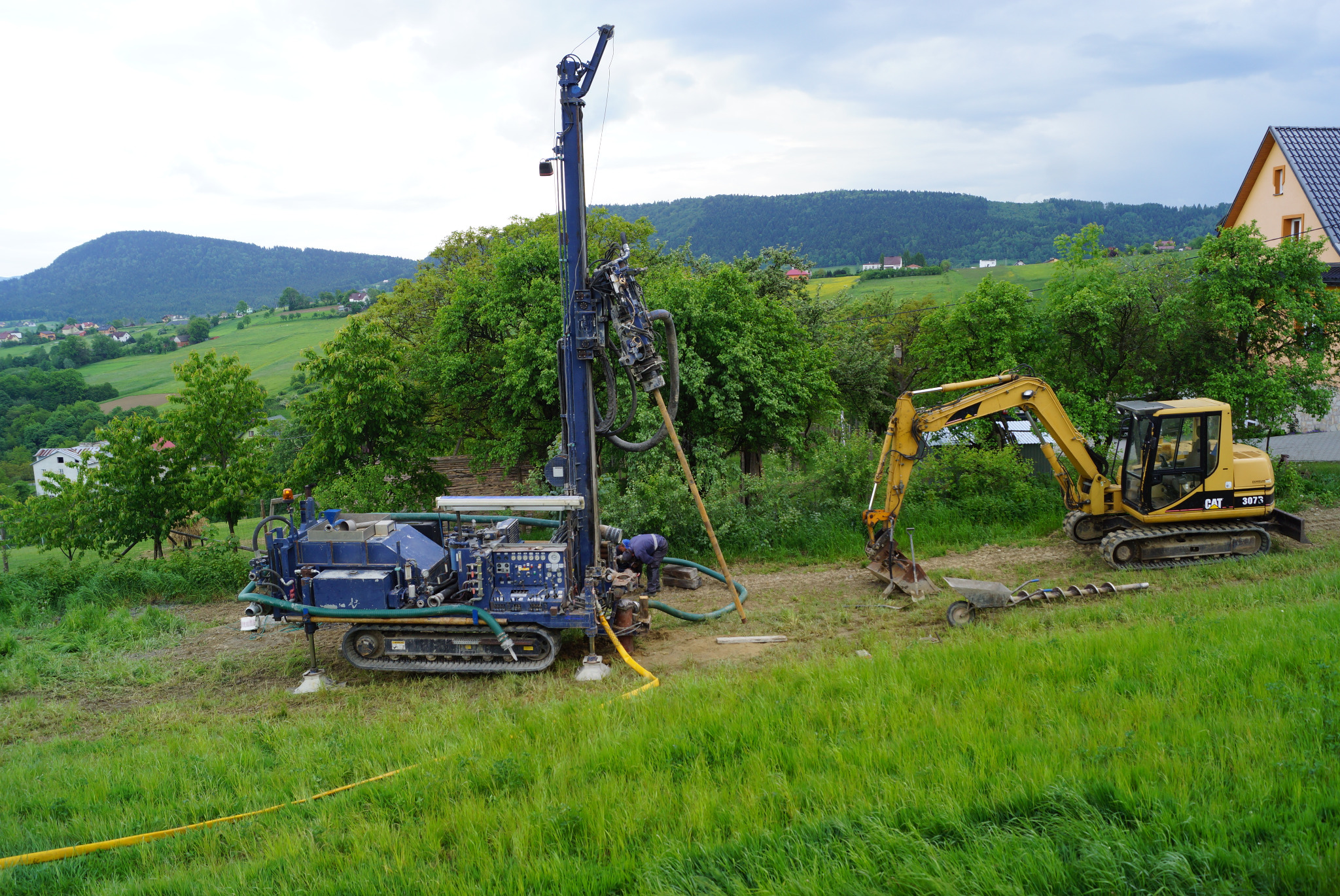 Niebieska wiertnica na gąsienicach podczas pracy na zielonej łące, obok żółta koparka CAT 307.3 i pracownik w kombinezonie, w tle pagórkowaty krajobraz i dom