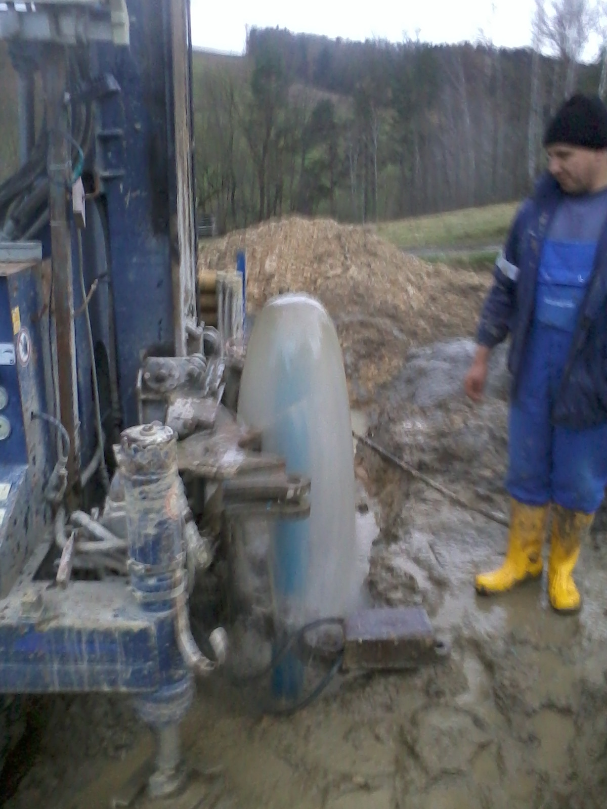 Wiertnica w trakcie pracy, woda tryska spod wiertła, obok stoi pracownik w żółtych gumowcach na rozmokniętym terenie, w tle widać drzewa.