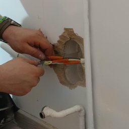 USŁUGI BUDOWLANO-ELEKTRYCZNE Damian Lipieński - Elektryk łączy przewody w ścianie z widocznym otworem, obok rury centralnego ogrzewania.