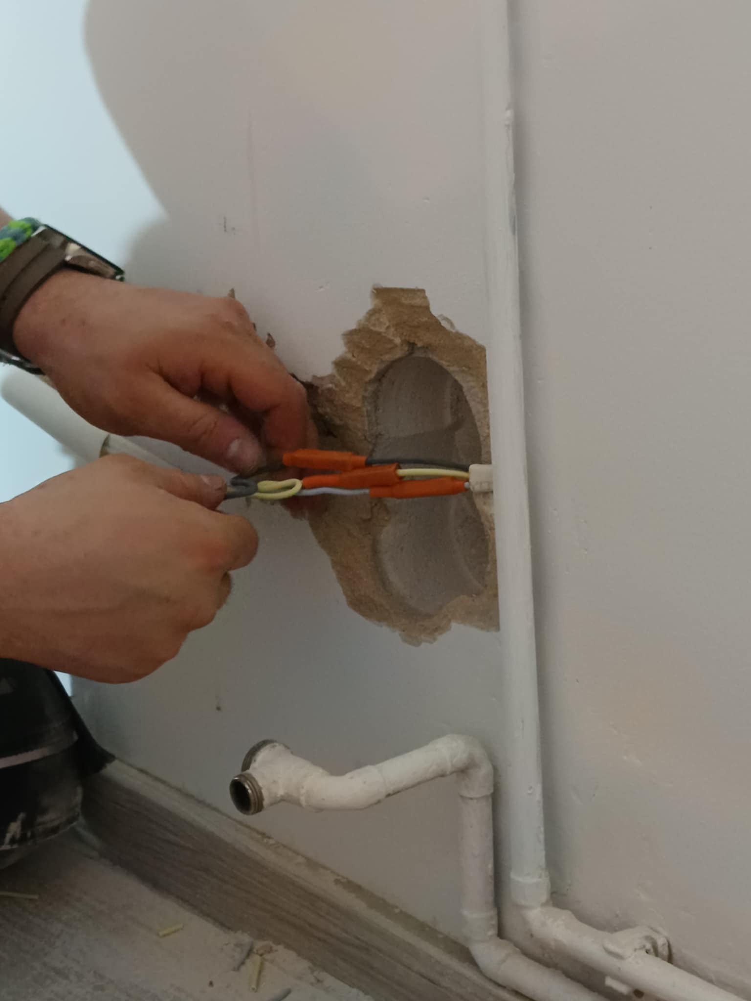 Elektryk łączy przewody w ścianie z widocznym otworem, obok rury centralnego ogrzewania.