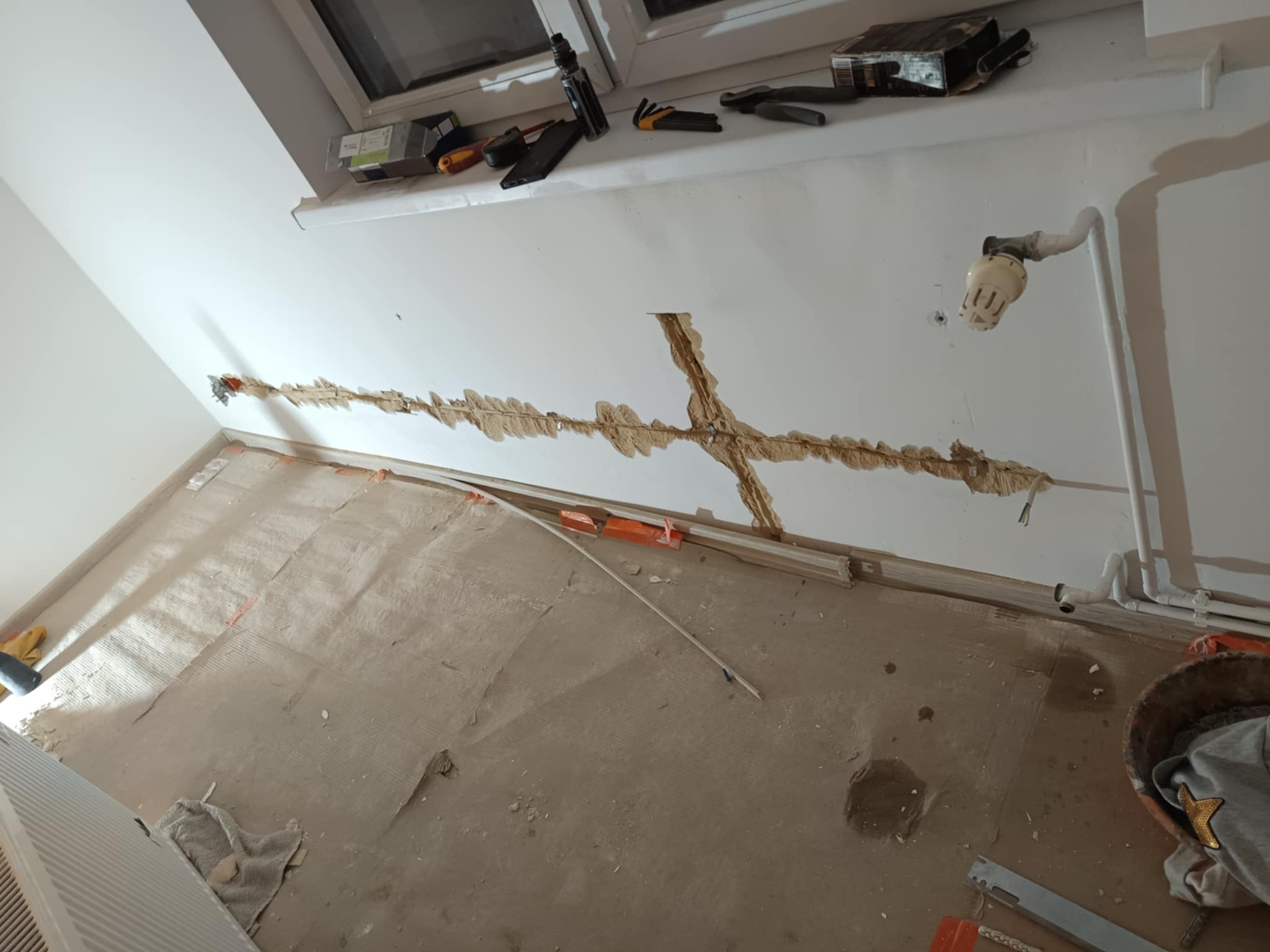 Przygotowanie ściany pod instalację elektryczną: wyżłobione bruzdy w ścianie pod kable, widoczne narzędzia na parapecie, rury grzewcze przy ścianie, podłoga zabezpieczona kartonem.