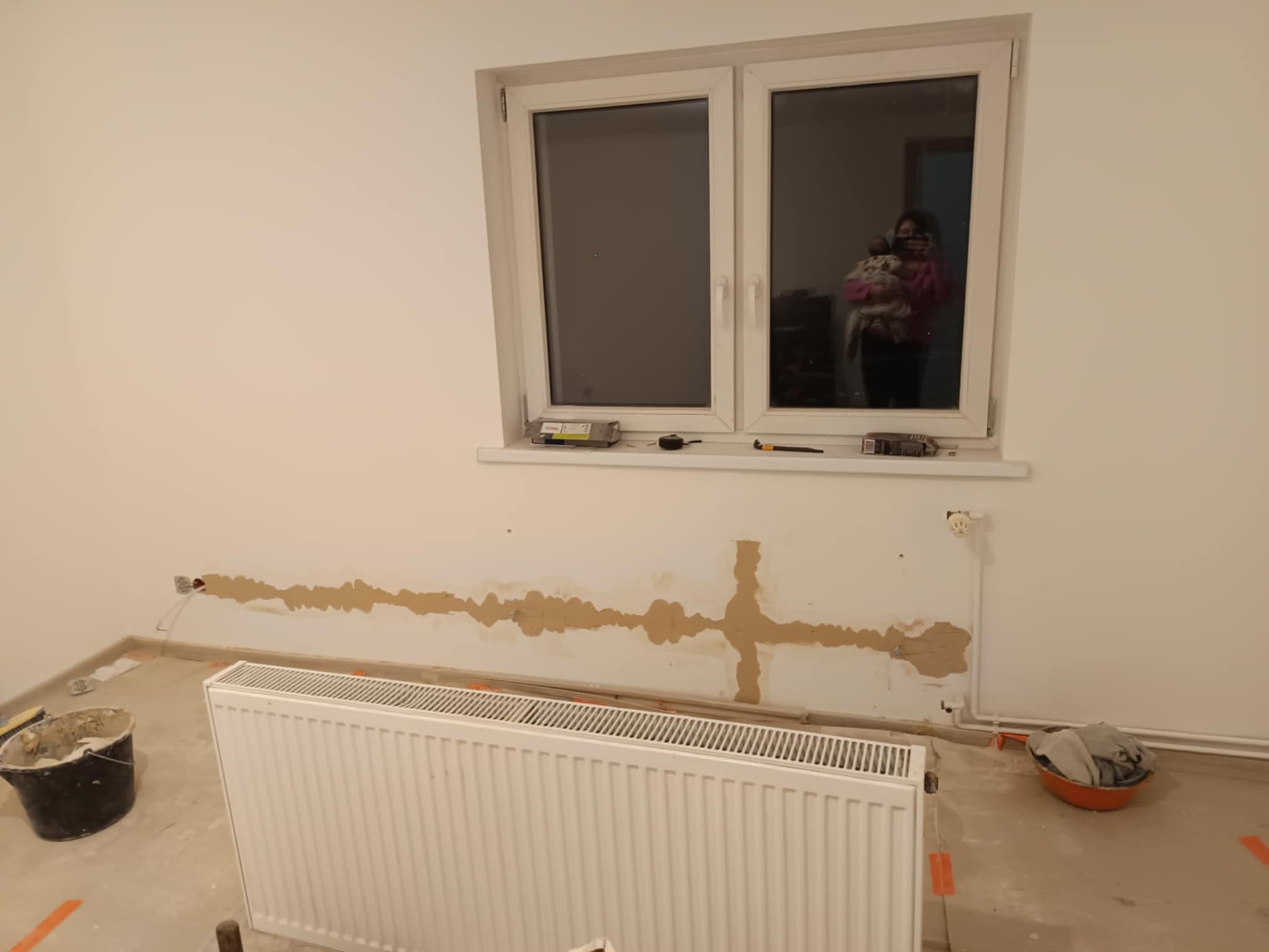 Przygotowanie ściany pod instalację elektryczną i grzejnika: wyfrezowane bruzdy pod kable, widoczny grzejnik, narzędzia na parapecie okiennym, odbicie osoby z dzieckiem w szybie.
