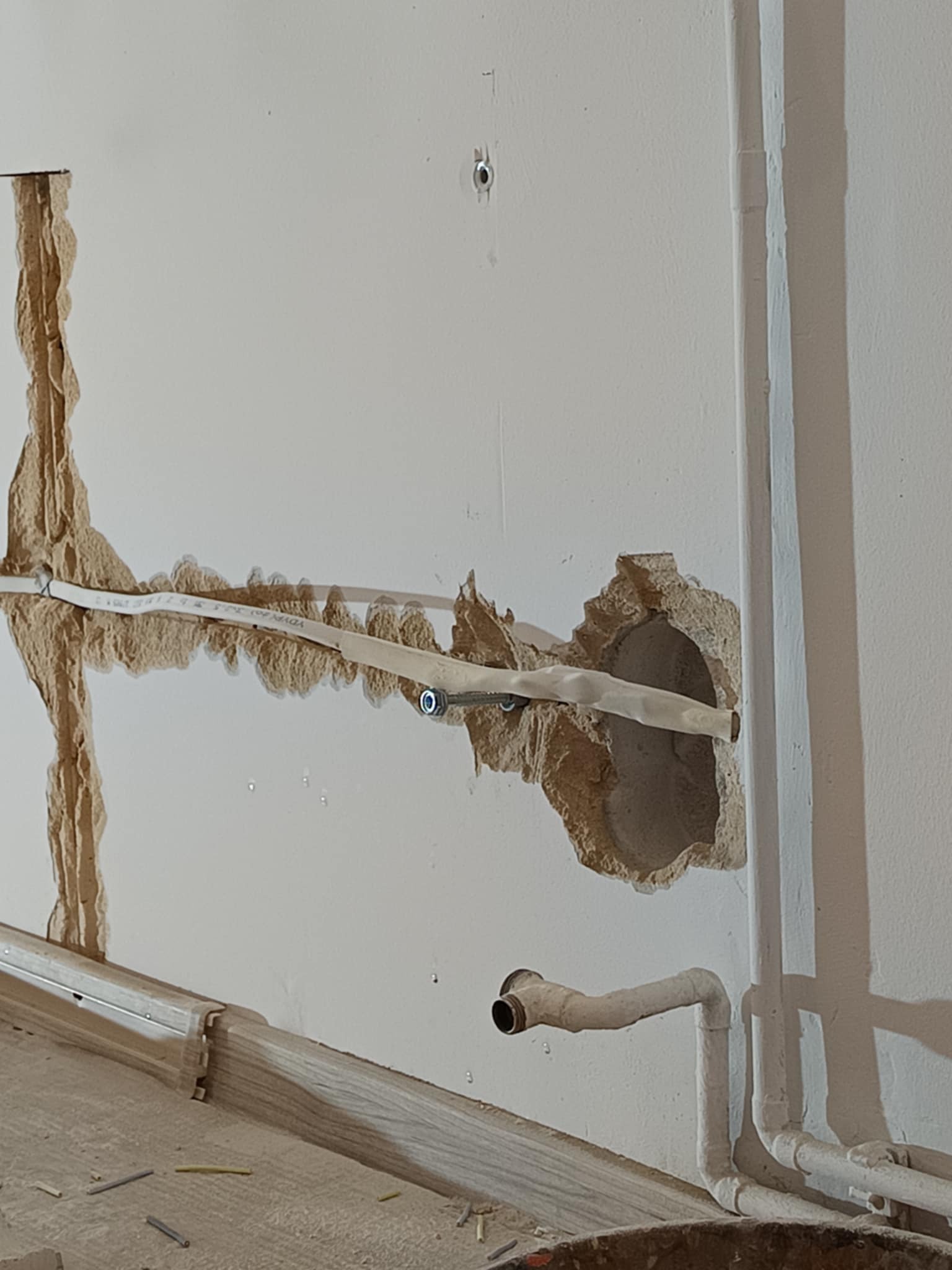 Biały kabel elektryczny prowadzony w bruździe w ścianie z widocznymi uszkodzeniami tynku. Dodatkowo widoczne rury instalacji sanitarnej.