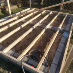 Konstrukcja drewniana szkieletowa na fundamencie z bloczków betonowych, widoczne przewody elektryczne i narzędzia, w tle siatka ogrodzeniowa.