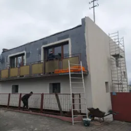 Ocieplanie elewacji dwupiętrowego budynku z balkonem, widoczne rusztowanie, drabiny, styropianowe płyty i pracownicy w trakcie montażu, pochmurne niebo w tle.