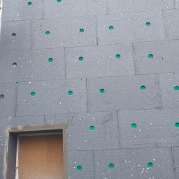 Mar bud - Szare płyty styropianowe z zielonymi kołkami mocowane do ściany budynku, widoczny otwór okienny z butelką w środku, etap prac.