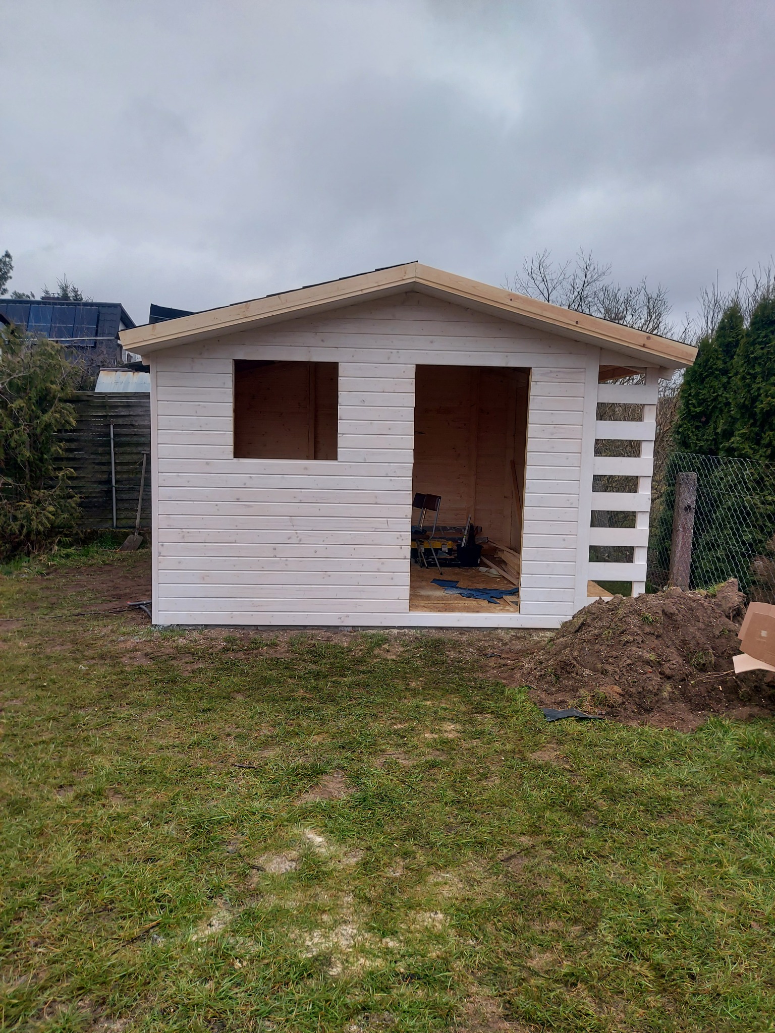 Drewniana konstrukcja domku letniskowego w trakcie budowy, widoczne otwory okienne i drzwiowe, na zewnątrz sterta ziemi i narzędzia.