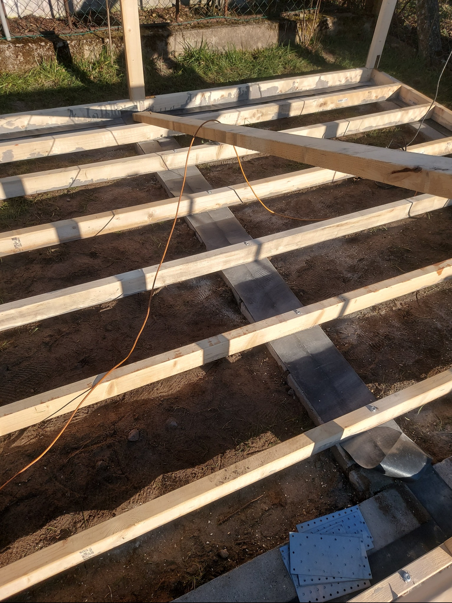 Konstrukcja drewniana pod taras na gruncie, widoczne legary, podkłady z papy, kabel zasilający i metalowe łączniki.