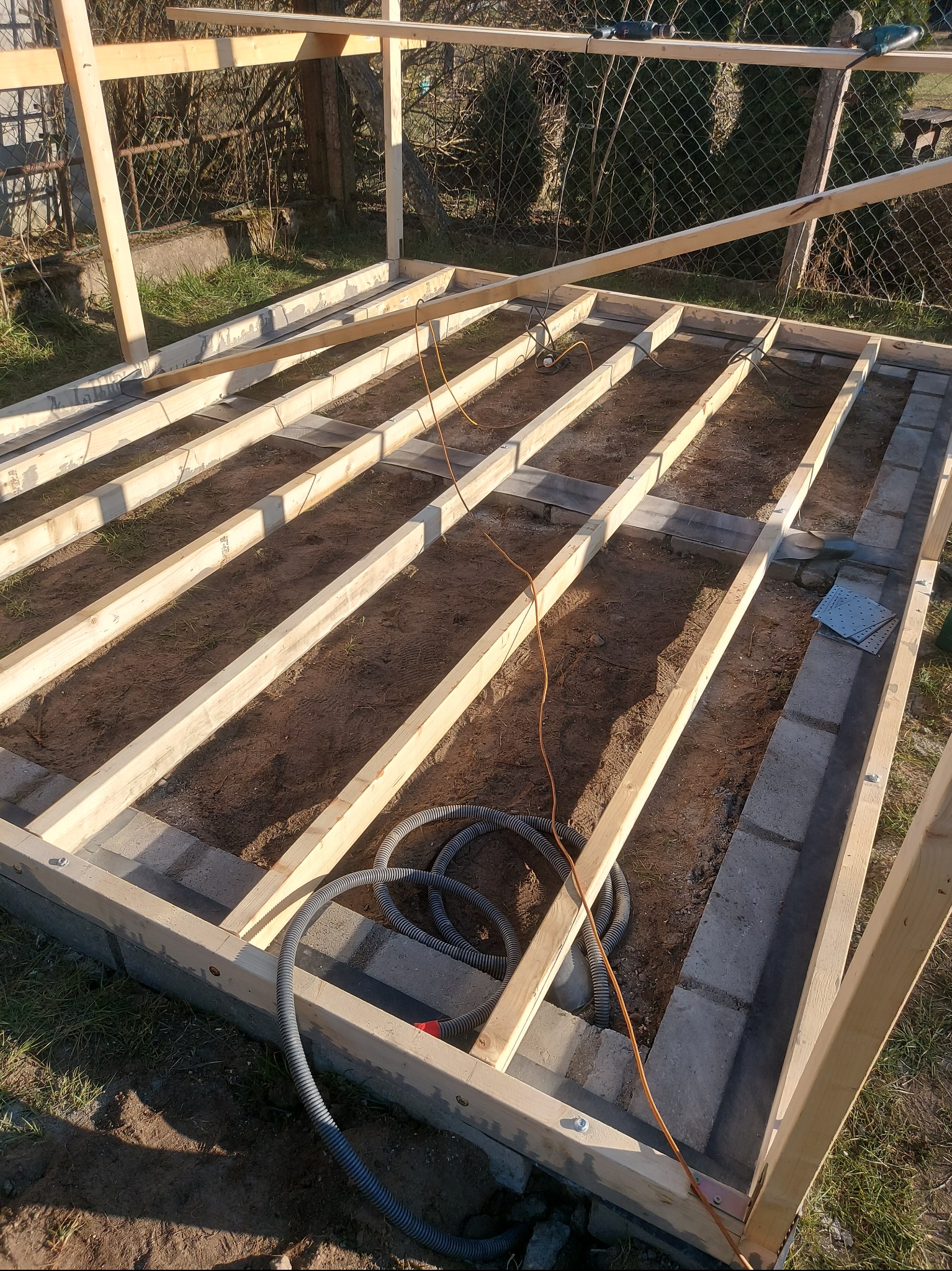 Konstrukcja drewniana szkieletowa na fundamencie z bloczków betonowych, widoczne przewody elektryczne i narzędzia, w tle siatka ogrodzeniowa.