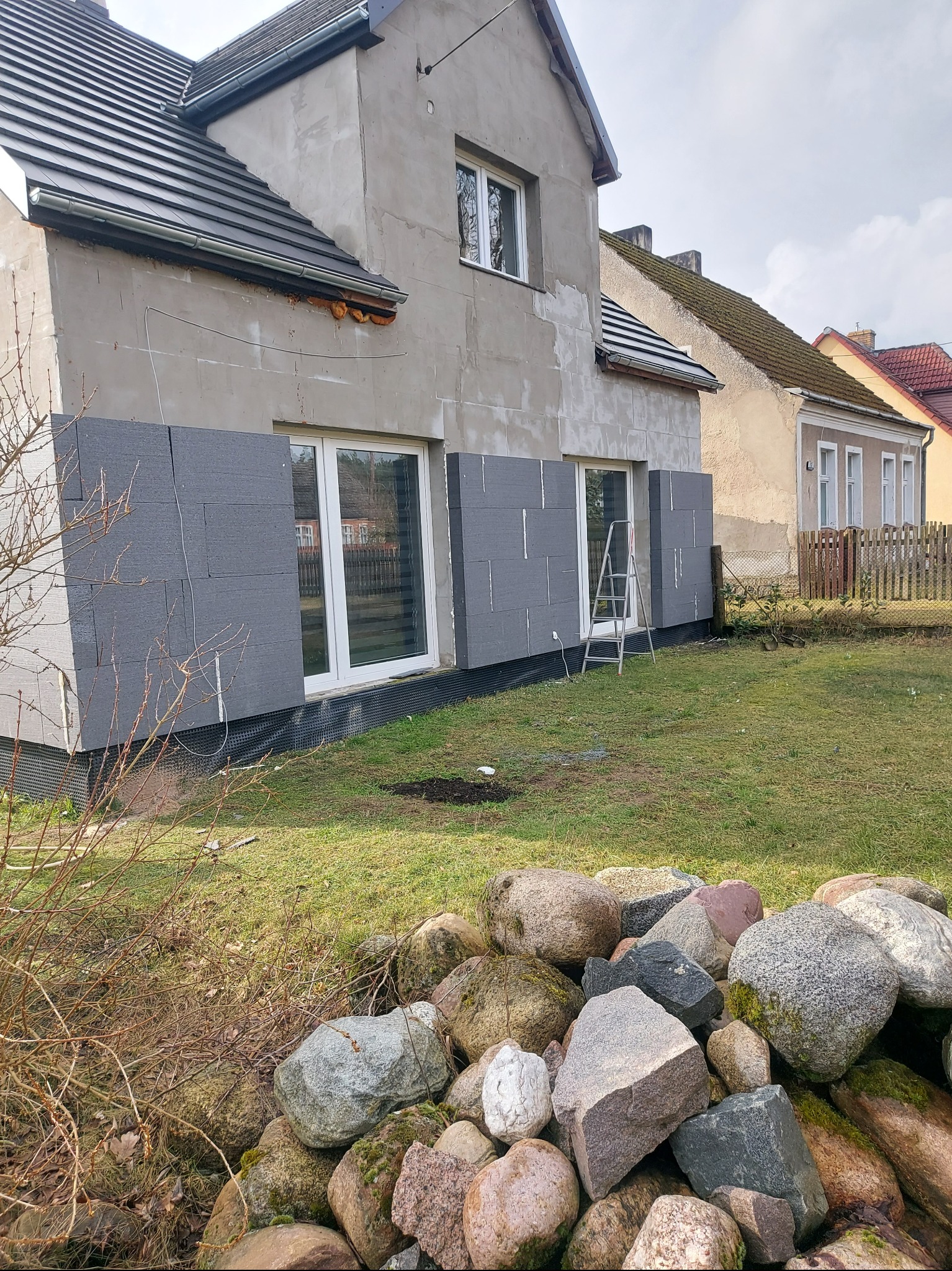 Dom w trakcie docieplania elewacji styropianem grafitowym, widoczne fragmenty ocieplenia przy oknach, drabina oparta o ścianę, na pierwszym planie kamienie.