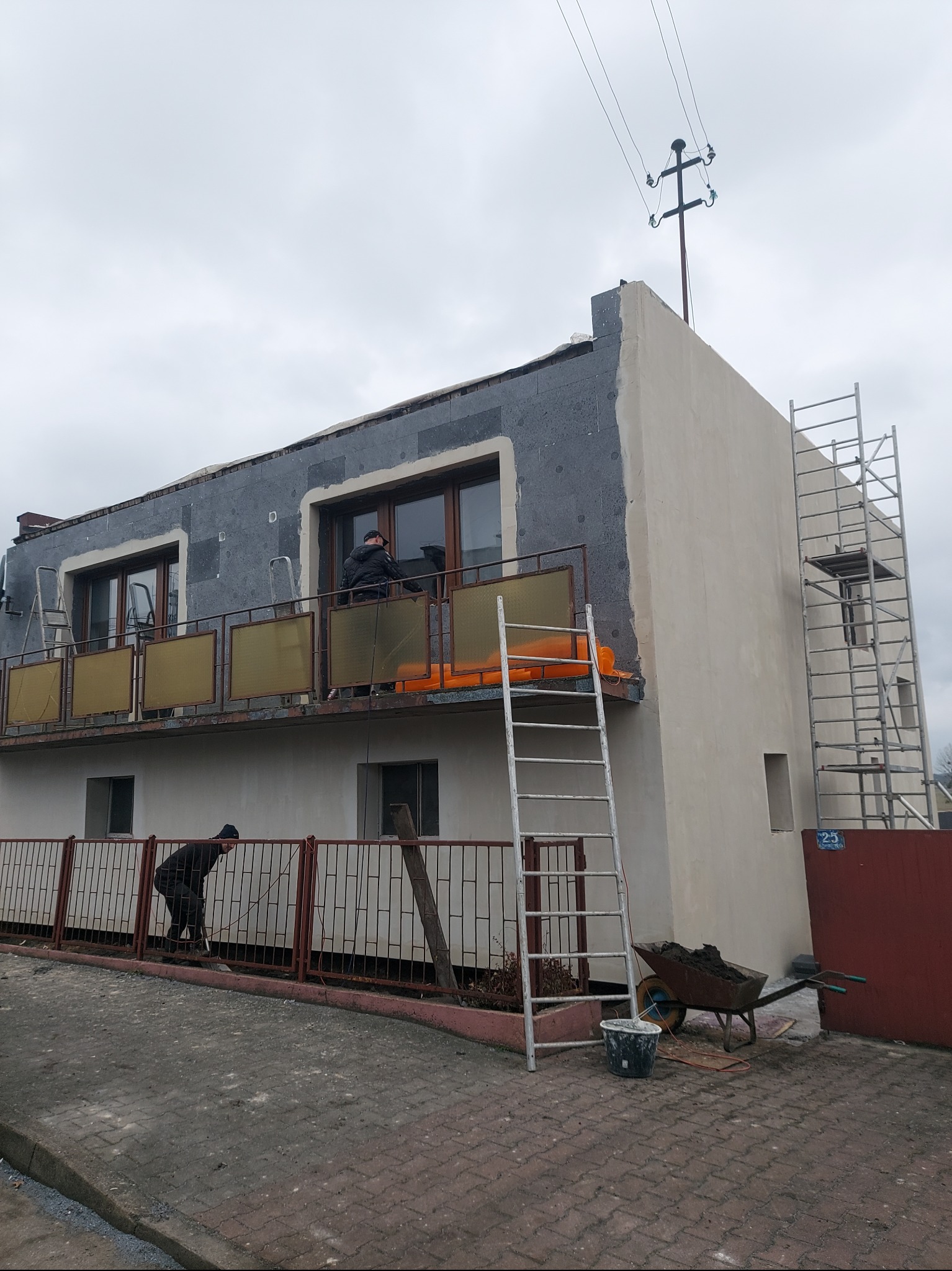 Ocieplanie elewacji dwupiętrowego budynku z balkonem, widoczne rusztowanie, drabiny, styropianowe płyty i pracownicy w trakcie montażu, pochmurne niebo w tle.