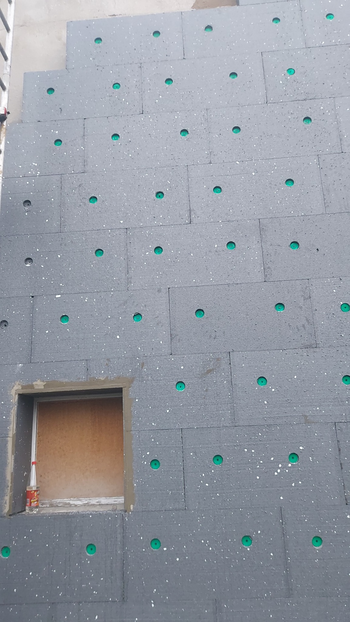 Szare płyty styropianowe z zielonymi kołkami mocowane do ściany budynku, widoczny otwór okienny z butelką w środku, etap prac.
