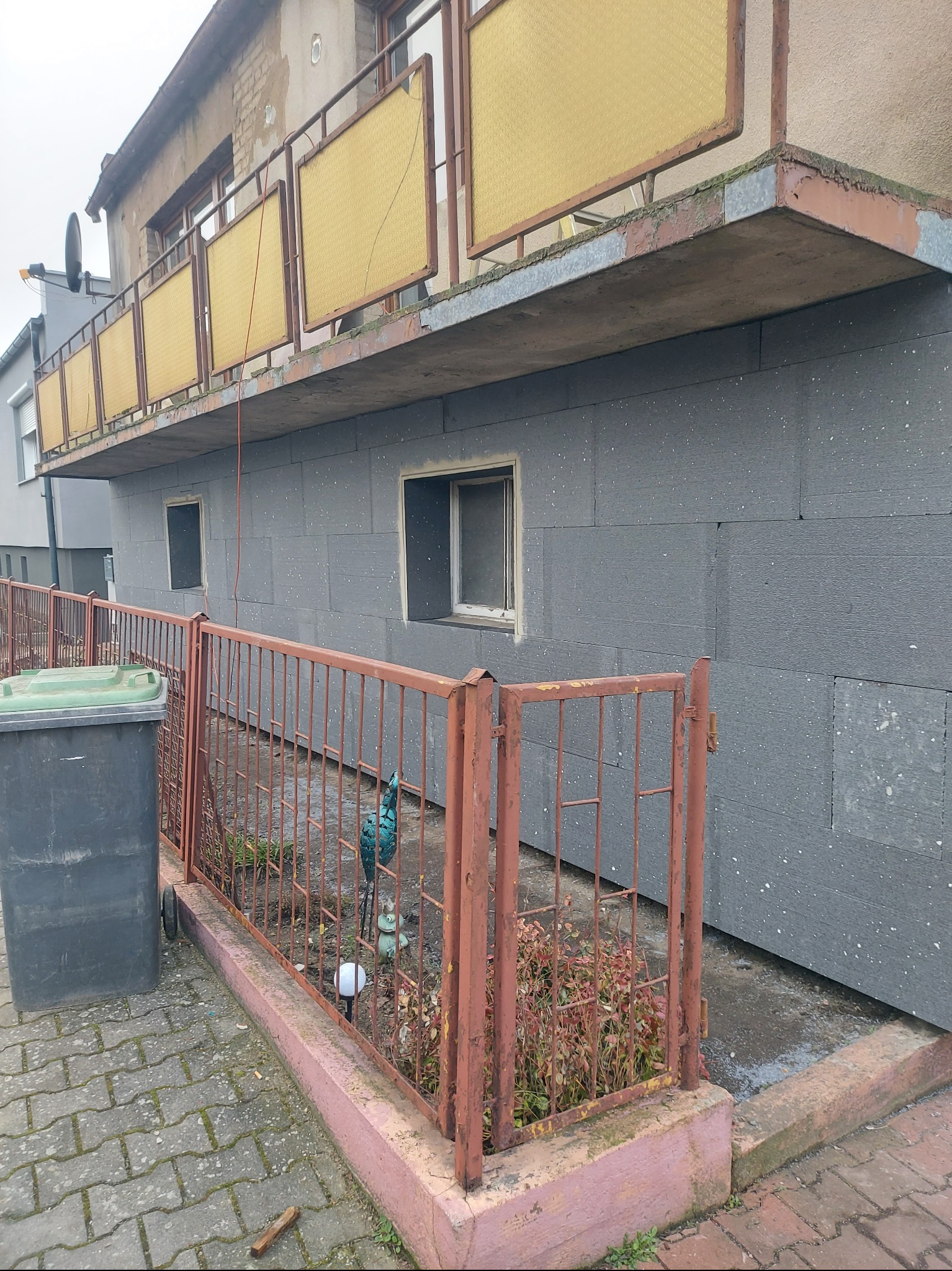 Budynek z ocieploną elewacją z szarych płyt styropianowych, widoczna stara, zardzewiała metalowa balustrada i śmietnik. Na balkonie żółte osłony z wzorem.